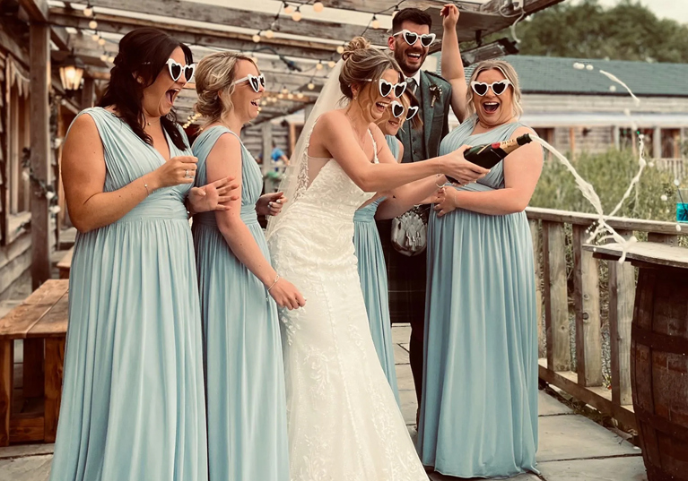 Bride, groom and bridesmaids wearing love heart shaped sunglasses pop bottle of champagne